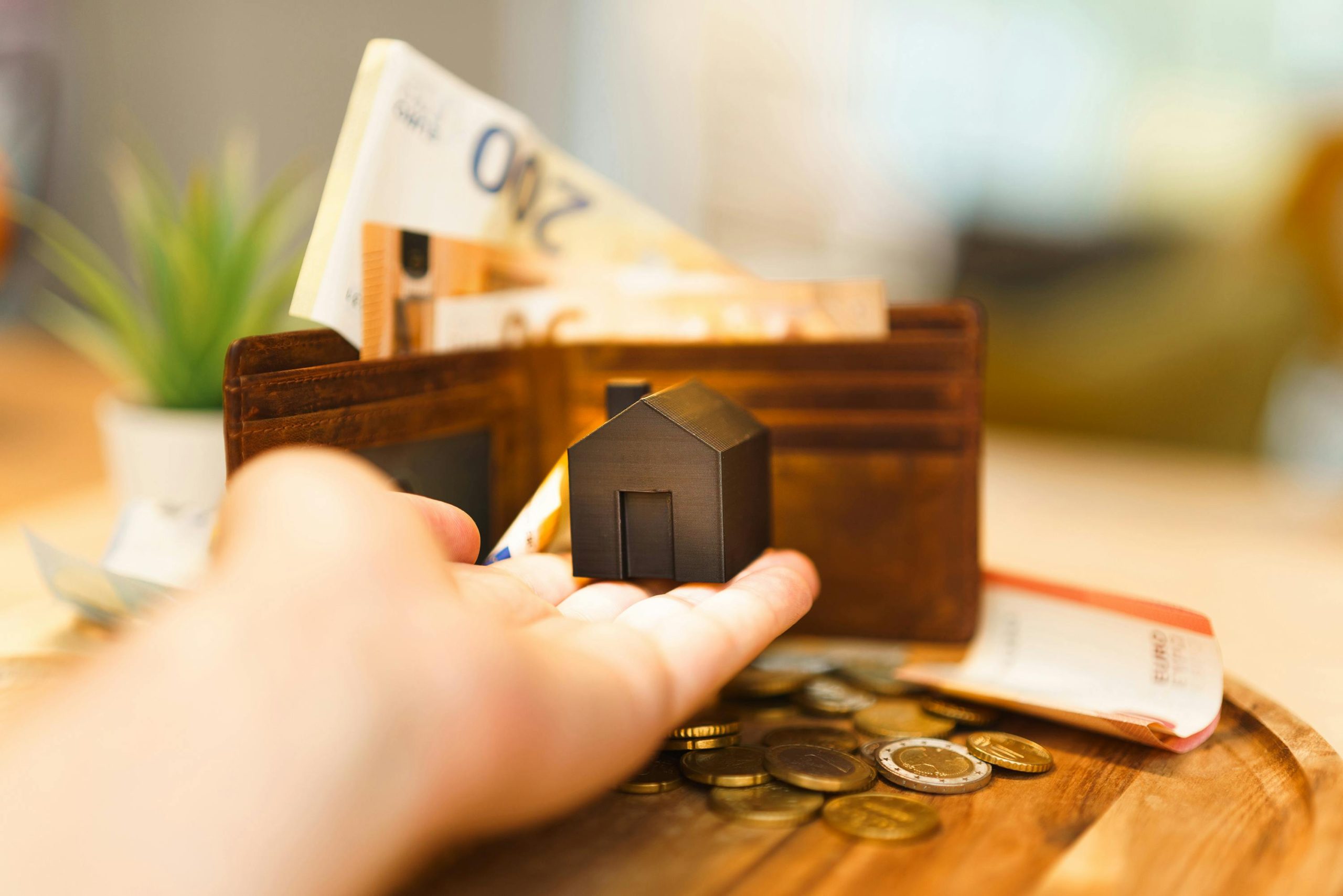 small house model resting on the palm of a hand, with a wallet and coins in the background